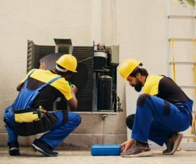 Two men in blue overalls troubleshooting AC problems while repairing an air conditioner in a well-lit indoor environment.
