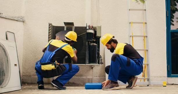 Two men in blue overalls troubleshooting AC problems while repairing an air conditioner in a well-lit indoor environment.