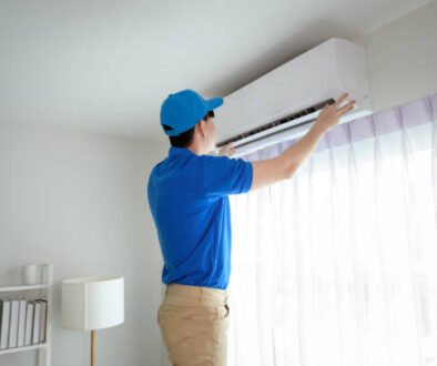 A man wearing a blue shirt and cap repairs an air conditioner, focused on his task.