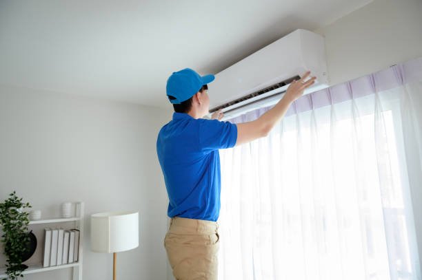A man wearing a blue shirt and cap repairs an air conditioner, focused on his task.