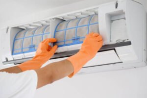 A person in orange gloves repairs an air conditioner, focusing on the unit with tools in hand.