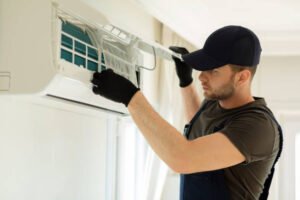 A technician wearing a hat and gloves performs annual AC maintenance, inspecting and repairing the air conditioner's internal components.
