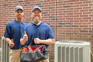 Two HVAC technicians in blue shirts hold tools and give thumbs up, smiling confidently at the camera after helping homeowners choose the right HVAC system for their South Florida home.
