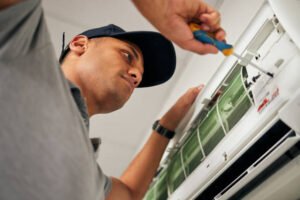 A man is repairing an air conditioner, focused on the unit with tools in hand, to improve indoor air quality in the home.