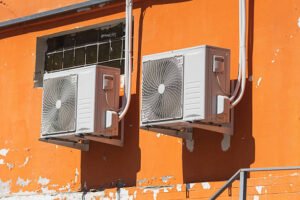 Two air conditioners mounted on a wall in front of an orange building, showcasing their AC compressor units.