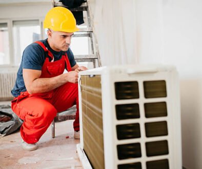 A man wearing overalls and a hard hat is engaged in maintenance work on an Ac compressor.