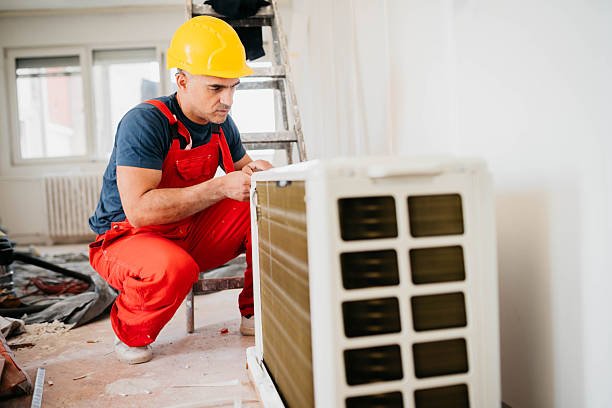 Worker works on broken air conditioner A man wearing overalls and a hard hat is engaged in maintenance work on an Ac compressor.