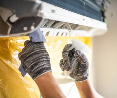 A person in gloves and a mask cleans an AC unit, ensuring proper maintenance and hygiene.
