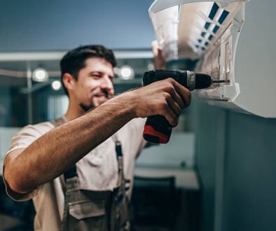 A man is using a drill to repair an air conditioner, demonstrating home maintenance skills.