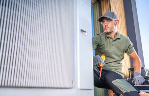 A man in a green shirt and hat repairs an air conditioner, representing HVAC and air conditioning contractors at work.
