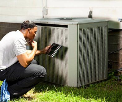 A man in work attire kneels next to an air conditioner, conducting HVAC maintenance.