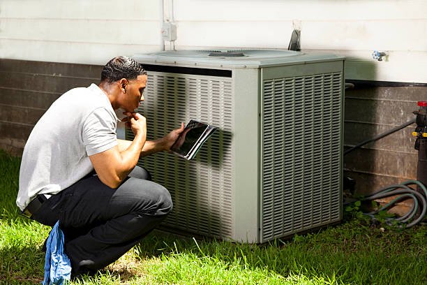A man in work attire kneels next to an air conditioner, conducting HVAC maintenance.