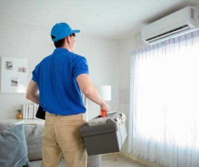 A man in a blue shirt carries a box in front of a window, representing an AC repair service in action.