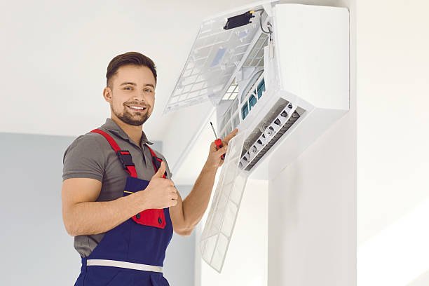 A man in overalls and an apron proudly holds a white air conditioner, promoting new air conditioning installation services.