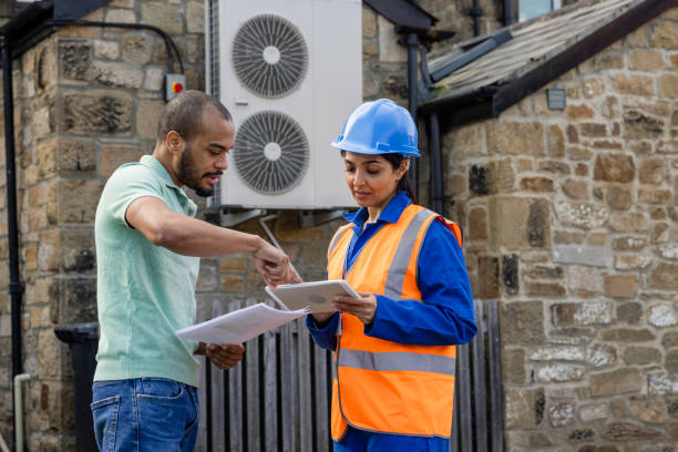 Two men in safety gear point at an air conditioner, engaged in a conversation about its installation process.
