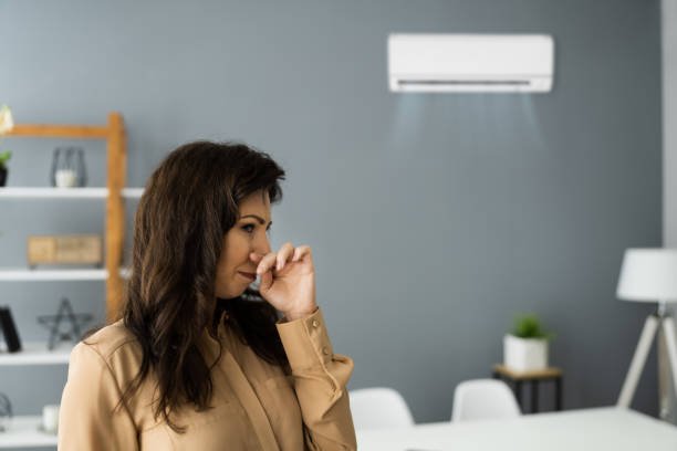 A woman stands near a wall air conditioner, looking concerned about a bad smell coming from the unit.