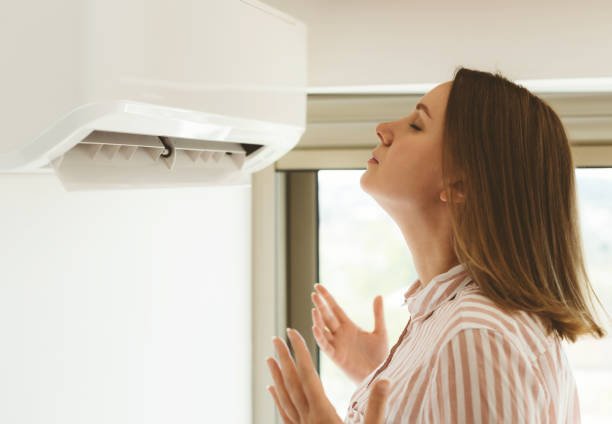 A woman stands near a wall air conditioner, looking concerned about a bad smell coming from the unit.