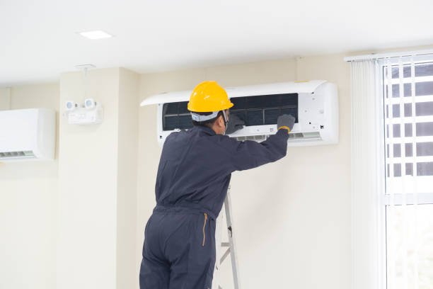 A man in a hard hat and overalls repairs an air conditioner during an installation process.