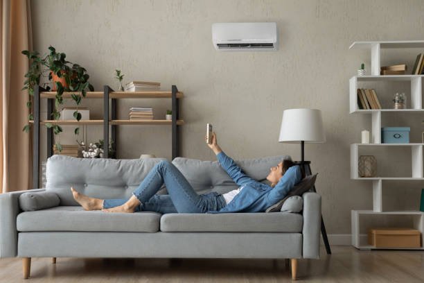 A woman relaxes on a couch, enjoying cool air from an air conditioner in a comfortable living room setting.