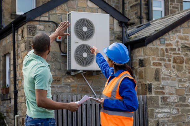 Two men in hard hats and vests point at an air conditioner during an installation discussion.