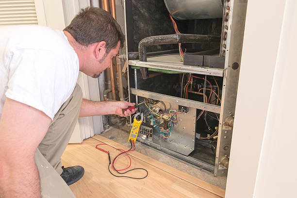 A man repairs a refrigerator in a residential HVAC service setting. Tools and parts are visible around him.
