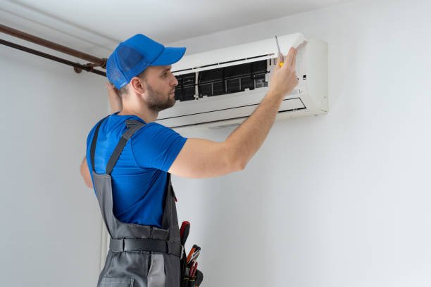 A man in overalls and a hat repairs an air conditioner, focused on fixing the unit.