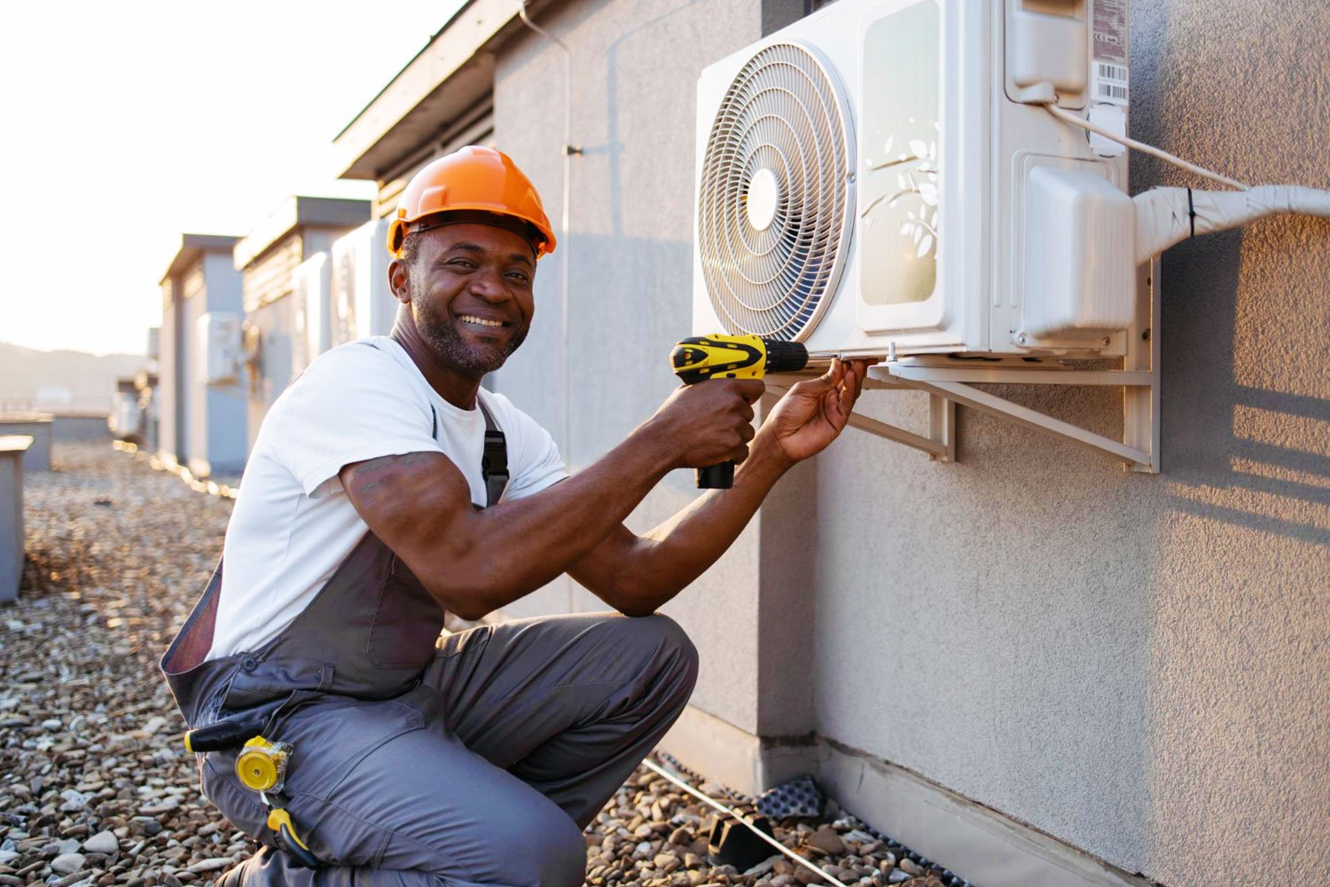 A smiling AC installation technician in an orange hard hat uses a power drill to install or repair an outdoor air conditioning unit on a rooftop.
