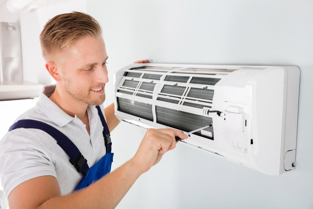 A male AC installation technician in blue overalls uses a screwdriver to service a wall-mounted mini-split air conditioner.