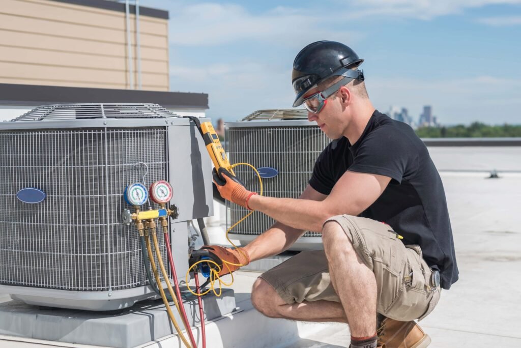 An HVAC technician in a hard hat and safety glasses kneels on a rooftop to inspect a large air conditioning unit using a digital multimeter and pressure gauges.