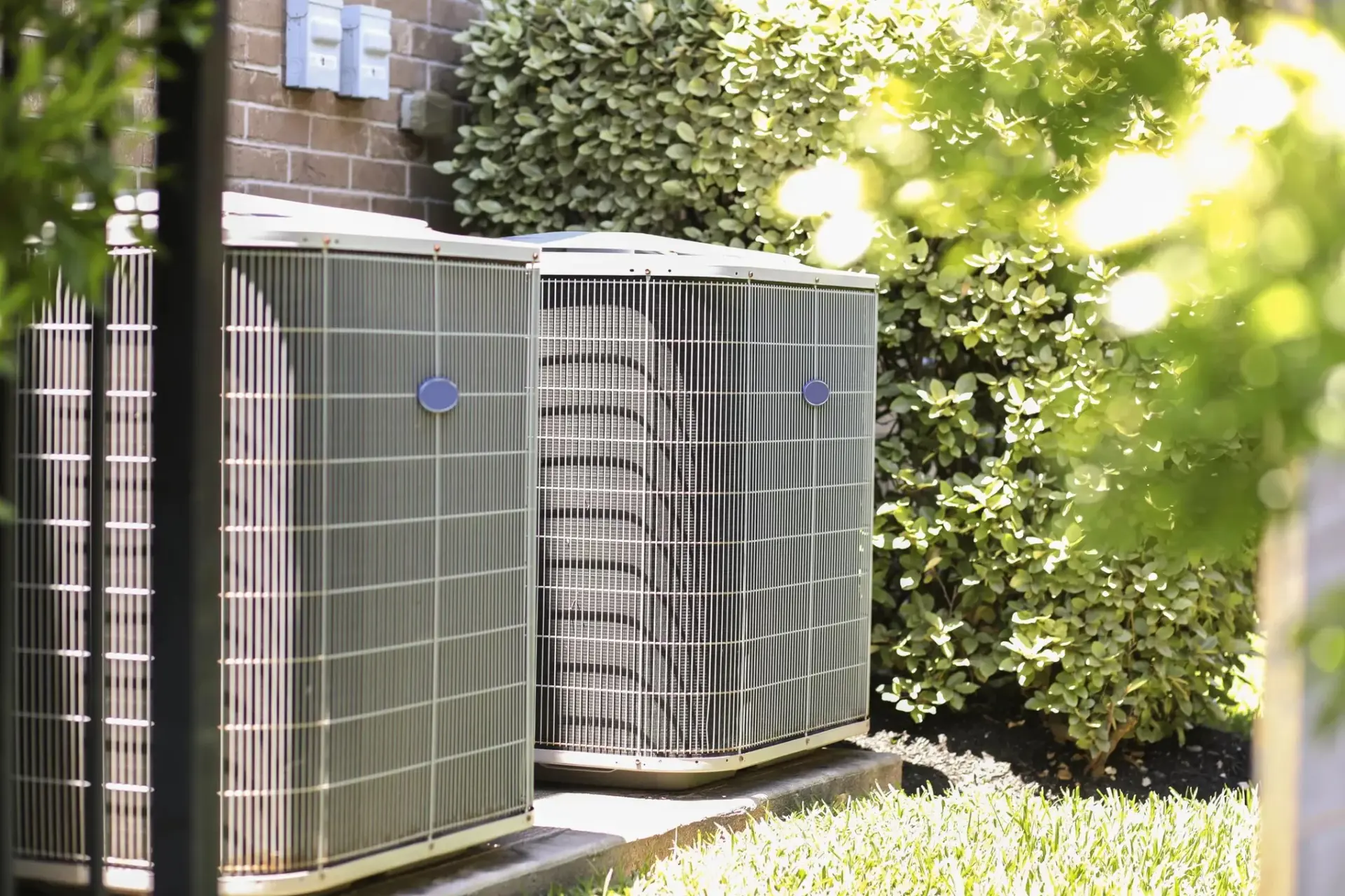 Two large outdoor air conditioning condenser units sit on concrete pads in a residential yard, bordered by a brick wall and green shrubbery.