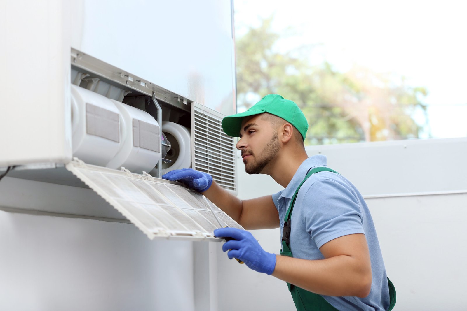 A man in a green shirt and blue gloves repairs an air conditioner, providing HVAC services.