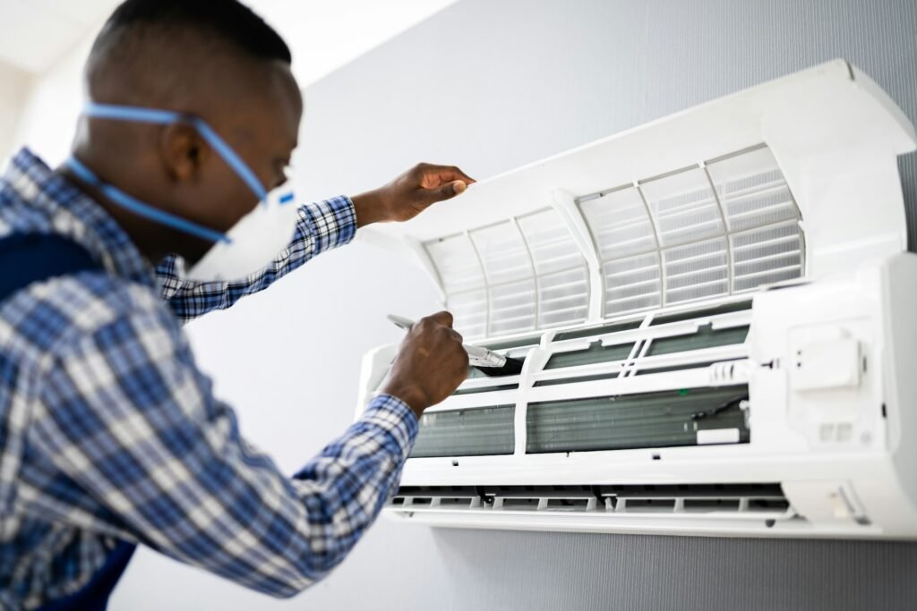 A man wearing a face mask and blue shirt repairs an air conditioner, focused on his task.