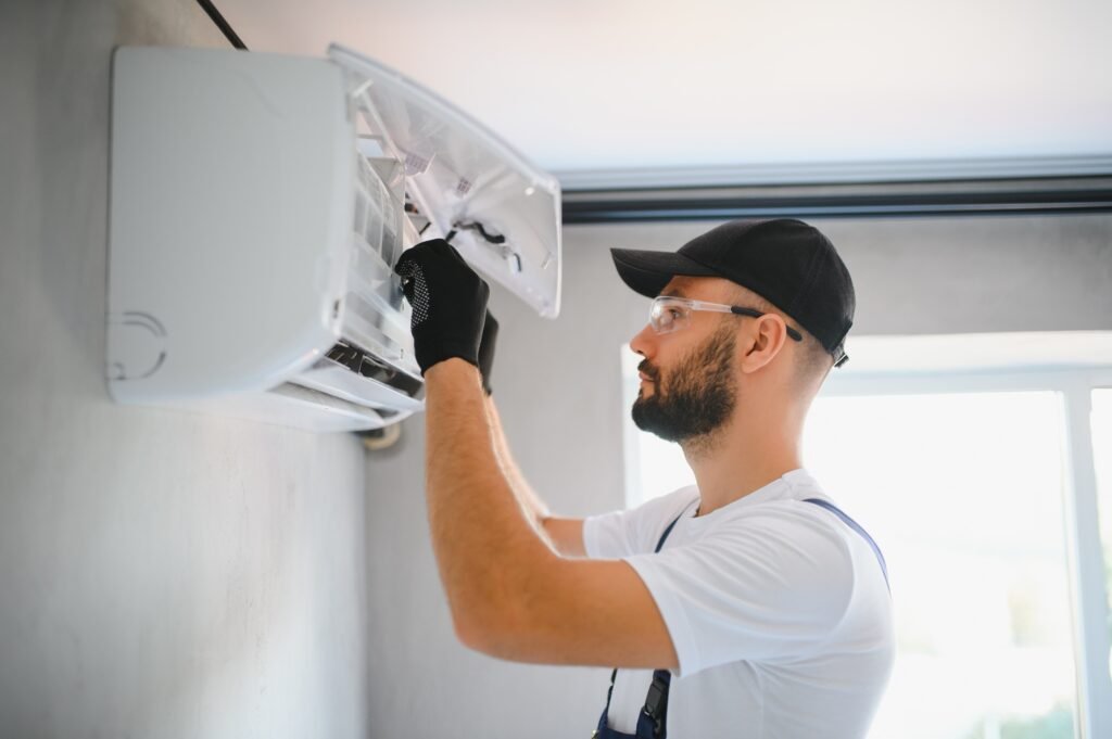 A man in a white shirt and hat repairing a residential air conditioner outdoors.
